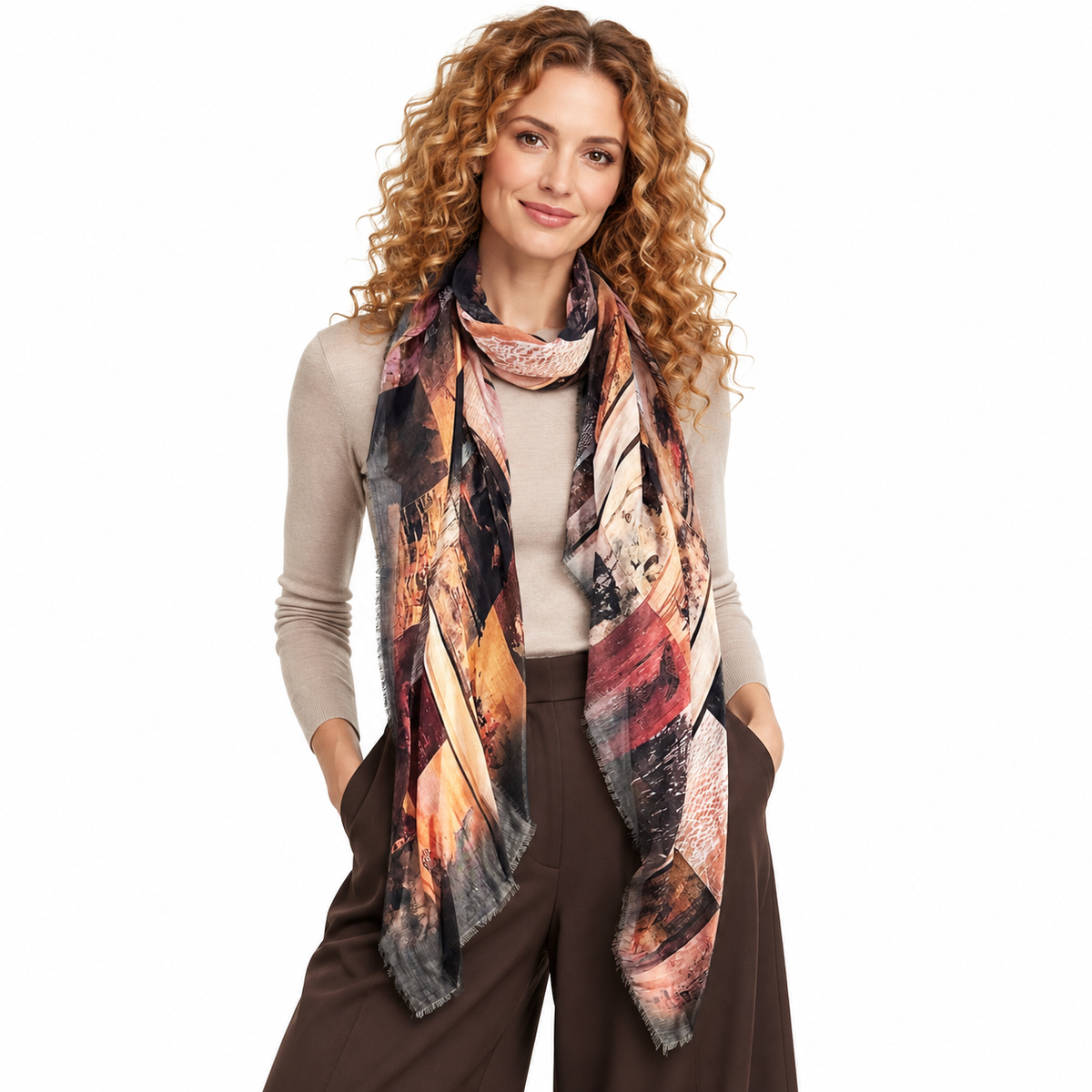 A woman with curly light brown hair models a long, lightweight scarf with an abstract geometric pattern in shades of orange, brown, beige, and black. She is wearing a beige long-sleeve top and dark brown wide-leg pants against a solid white background.