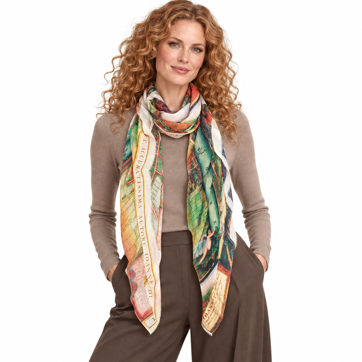 A woman with curly red hair models a multicolor square scarf with a vintage map and botanical print, styled over a taupe sweater and brown wide-leg trousers against a solid white background.