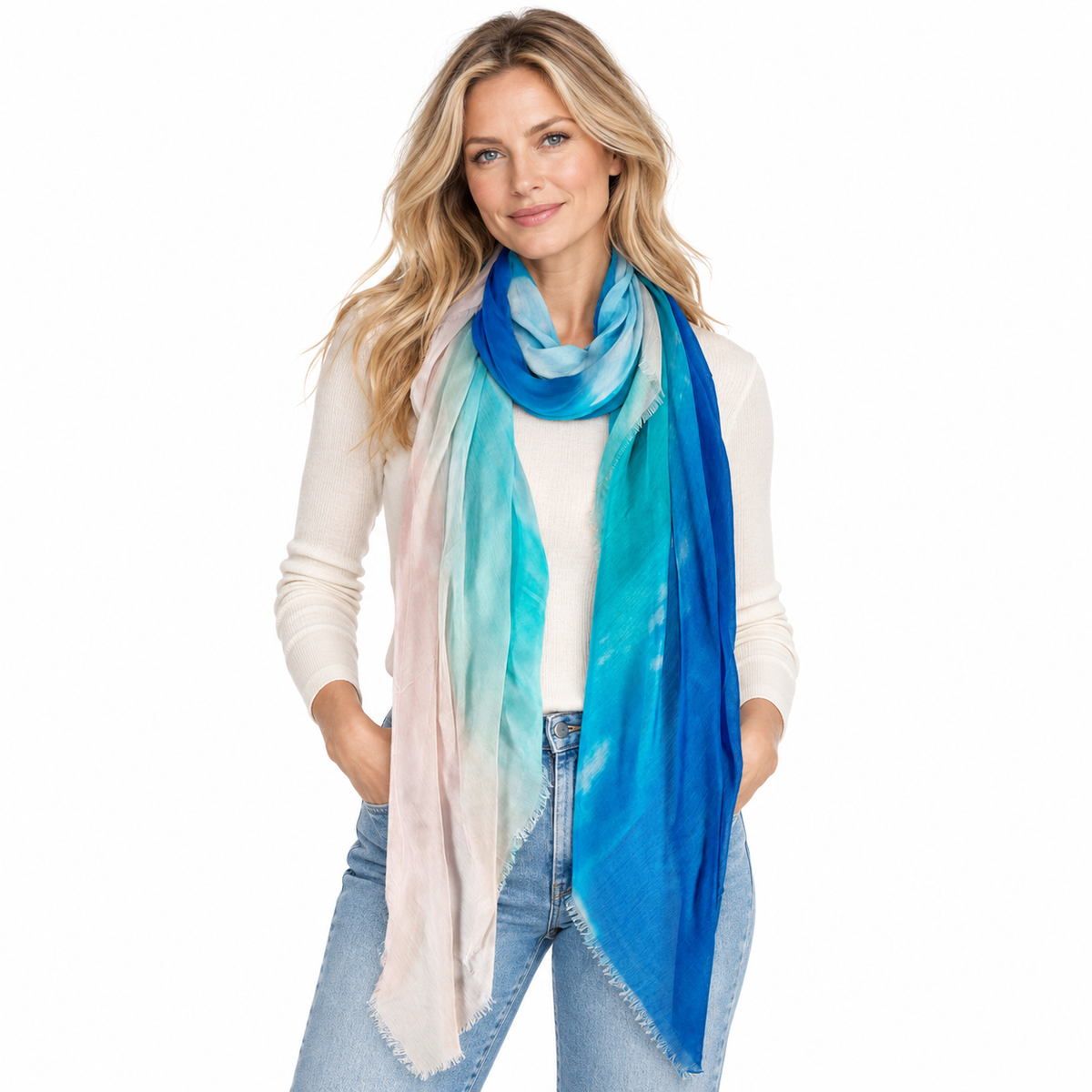 A woman modeling a lightweight scarf with a blue, teal, and pale pink tie-dye pattern, styled over a cream long-sleeve top and blue jeans against a plain white background.