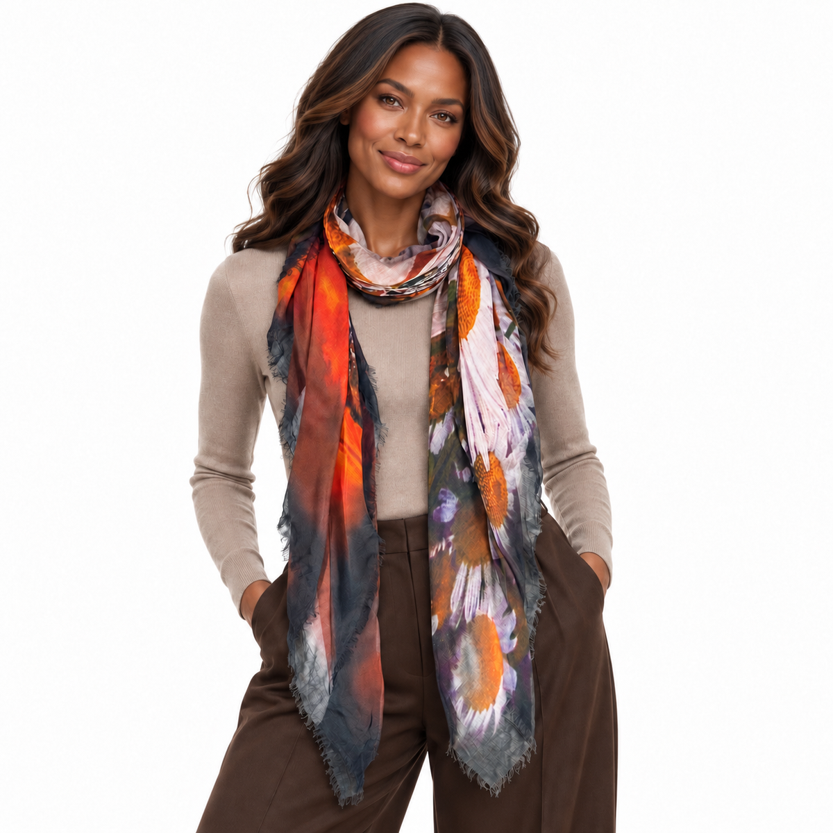 A woman modeling a lightweight, fringed scarf with an abstract floral pattern in orange, gray, purple, and white, worn over a beige sweater and brown pants against a white background.